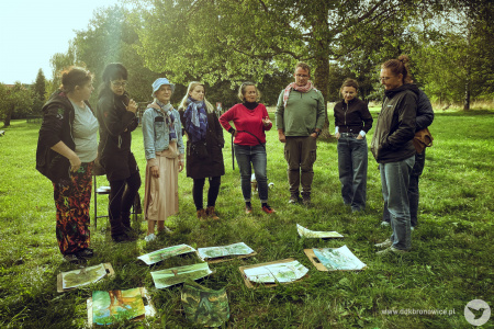 Plener malarski. Ośmioro uczestników. Park. Drzewa i krzewy. Grupa stoi w półkolu. Na środku leżą namalowane obrazy.