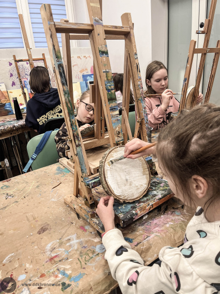 Grupa maluje swoje obrazy na drewnie. Na stołach rozciągnięty papier. Na pierwszym planie dziewczynka maluje białe tło.