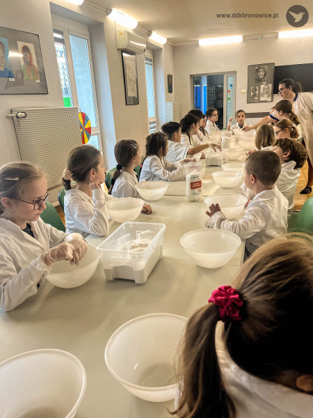 Na białym stole pojawiły się miski i dodatkowe słoiki. Grupa dzieci przy stole słucha pani prowadzącej.
