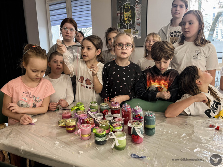 Dzieci pozują do zdjęcia ze swoimi kolorowymi świeczkami. Każdy słoiczek ma kolorową kokardkę. Słoiki leżą na stole.