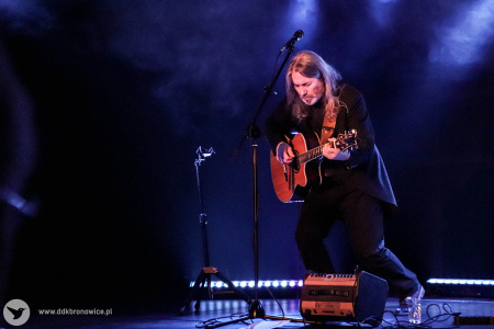 Artysta gra na gitarze na scenie. Za nim niebieskie światło.