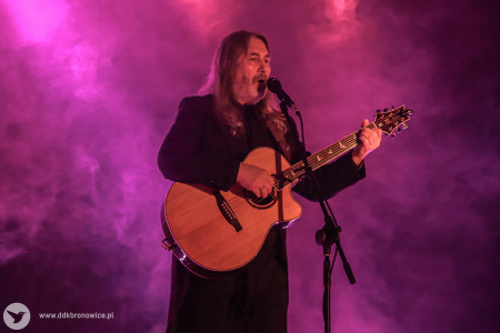 Jan Kondrak śpiewa do mikrofonu. Gra na gitarze. Świeci się czerwone światło. Na scenie unosi się dym.