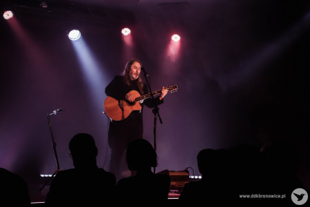 Na scenie muzyk wykonuje piosenkę. Widać zapalone reflektory. Na przodzie widać kontury publiczności.