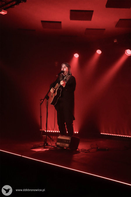 Jan Kondrak stoi na scenie. Śpiewa. Gra na gitarze. Zapalone czerwone światło.