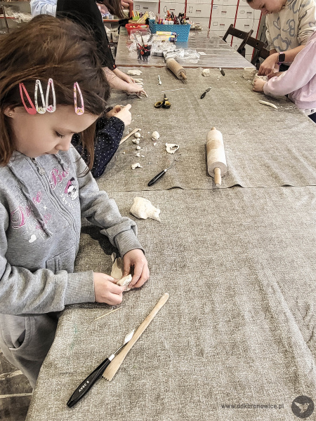 Dziewczynka w szarej bluzie stoi przy stole warsztatowym. Rękami nadaje kształt glinie. Tworzy mydelniczkę.