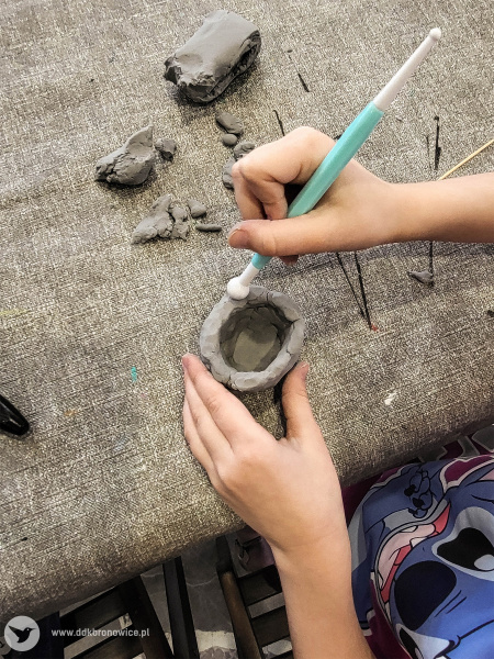 Dziewczynka robi swoje naczynie z gliny. Formuje je używając biało-niebieskiego prostego narzędzia.