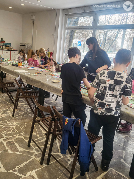 Sala warsztatowa. Przy stole stoją dzieci. Dziewczynki po lewej stronie siedzą. Dzieci malują swoje obrazy.