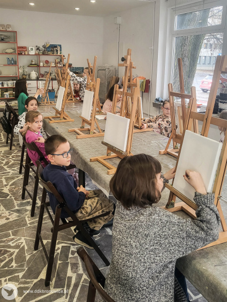 Dzieci siedzą przy stole warsztatowym. Na stole sztalugi i białe obrazy. Dzieci zaczynają malować.
