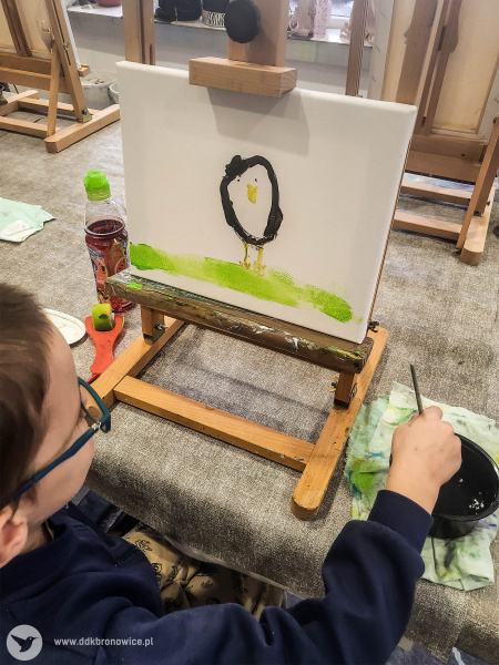 Chłopiec w granatowej bluzie maluje swój obraz na sztaludze. Na obrazie zielona trawa i mały okrągły ptak z żółtym dziobem.