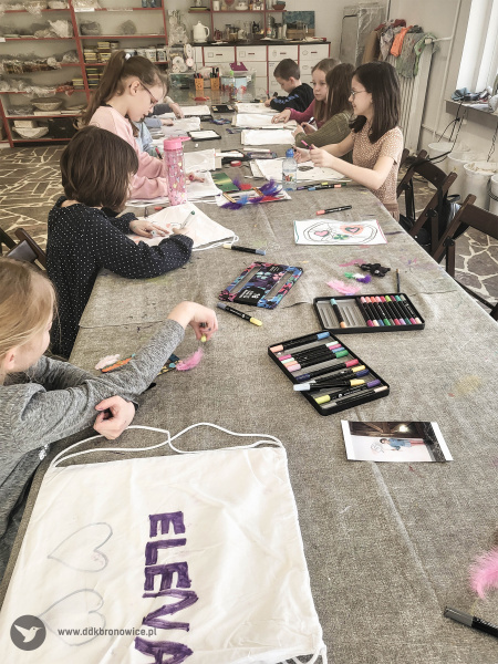 Dzieci przy stole warsztatowym tworzą swoje artystyczne obrazy. Na stole rozłożone markery.