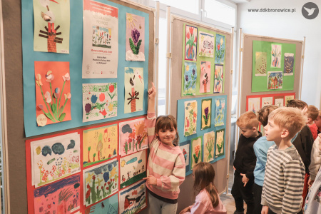 Dziewczynka w biało-różowej bluzie wskazuje swój obraz. Szeroko uśmiecha się. Jest szczęśliwa.