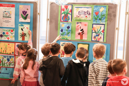 Dzieci oglądają obrazy. Obrazy przedstawiają kolorowe kwiaty i zielone drzewa. Dziewczynka wskazuje na swój obraz.