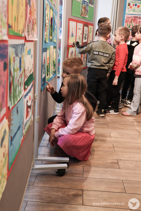 Dziewczynka wskazuje na swój obraz. Obraz jest nisko zawieszony. Dziewczynka, żeby go zobaczyć musi kucać.