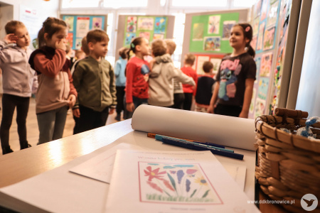 Na pierwszym planie rozłożone broszury dotyczące wystawy. Obok leża kredki. W tle dzieci oglądają obrazy.