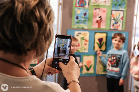 Kobieta robi telefonem zdjęcie chłopcu w niebieskiej bluzie. Chłopiec pozuje przy swoim obrazie.