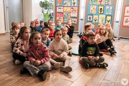 Dzieci siedzą na podłodze. Za nimi zawieszone obrazy.