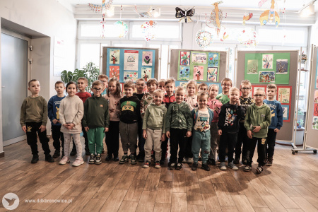 Cała grupa dzieci stoi i patrzy się w stronę aparatu. Uśmiechają się. Za nimi zawieszone obrazy.