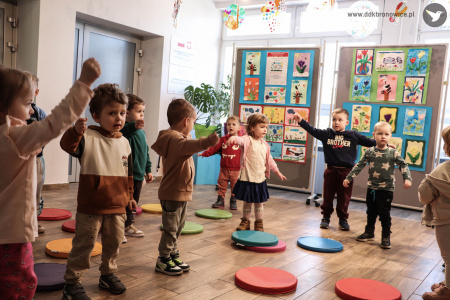 Dzieci stoją i mają podniesione ręce. Na podłodze rozłożone małe, okrągłe, kolorowe poduszki.