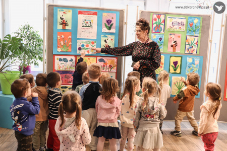 Dzieci stoją wokół nauczycielki. Kobieta ubrana w czarną sukienkę w kropki, wskazuje na jeden z obrazów.