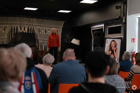 Na pierwszym planie widoczna publiczność. W tle, na scenie, występuje aktorka w pomarańczowym swetrze.