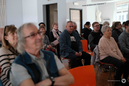 Osoby siedzące na widowni. Po lewej stronie starszy mężczyzna ubrany w koszule z krótkim rękawem uśmiecha się.