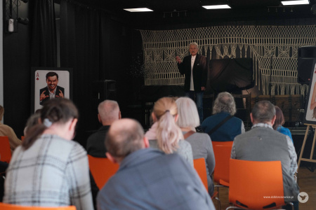 Na scenie aktor przemawia do publiczności. Ubrany jest w białą koszulkę i brązową marynarkę.