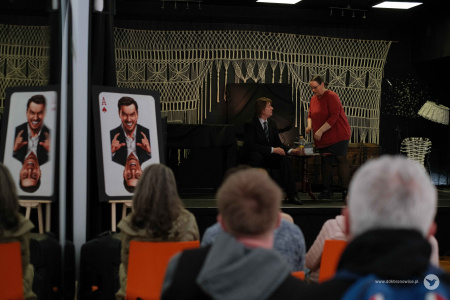 Na scenie aktor w granatowym garniturze siedzi przy stoliku. Rozmawia z aktorka w czerwonej bluzce. Aktorka stoi i pochyla się nad aktora.