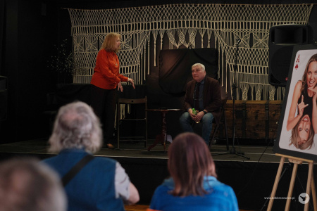 Aktor w brązowej marynarce i bordowym krawacie siedzi po prawej stronie sceny. Aktorka w pomarańczowym swetrze stoi obok niego. Rozmawiają.