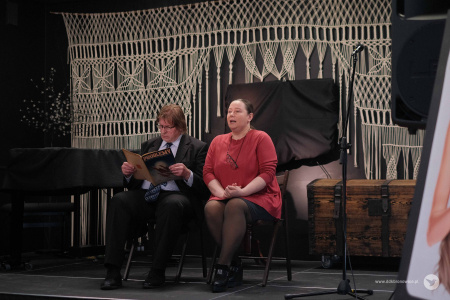 Na scenie aktor i aktorka. Aktor siedzi po prawej stronie i ubrany jest w granatowy garnitur z krawatem. Po prawej stronie siedzi aktorka w czerwonej bluzce. Między nimi stolik. Aktor czyta gazetę.