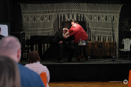 Aktorka w czerwonej bluzce pomaga rozpiąć  marynarkę aktorowi. Aktor siedzi i trzyma się za głowę.