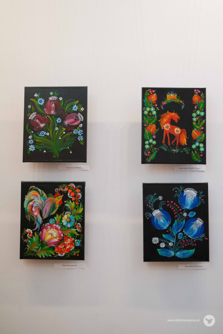 Na białej ścianie zawieszone cztery obrazy. Wszystkie mają czarne tło. Przedstawiają motywy kwiatowe i roślinne. Przeważają kolory zielone i czerwone. Obraz w dolnym prawym rogu jest niebieski.