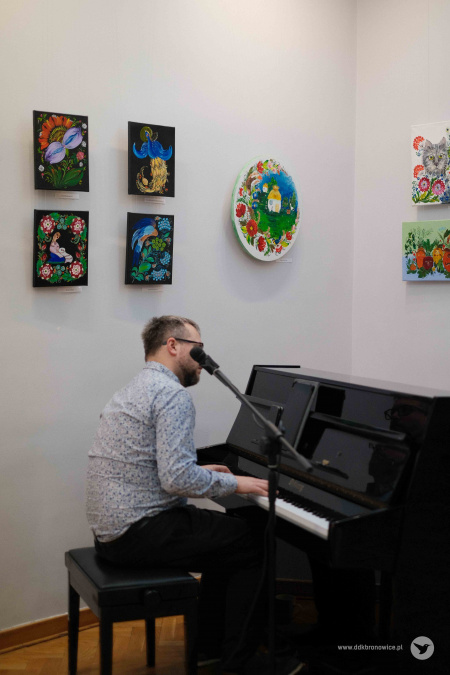 Mężczyzna w biało-niebieskiej koszuli gra na pianie. W tle kolorowe obrazy z motywami roślinnymi i zwierzęcymi.