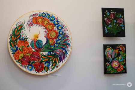 Po lewej stronie charakterystyczny, okrągły obraz przedstawiający koguta. Wokół zwierzęcia kwiaty i rośliny. Obok okrągłego obrazu zawieszone są dwa mniejsze kwadratowe z czarnym tłem.