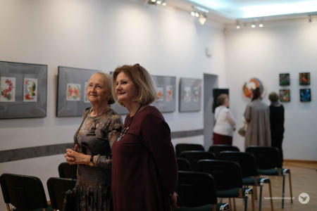 Dyrektor dzielnicowego domu kultury Bronowice rozmawia ze starszą kobietą ubraną w szara bluzkę. Podziwiają obrazy i uśmiechają się.