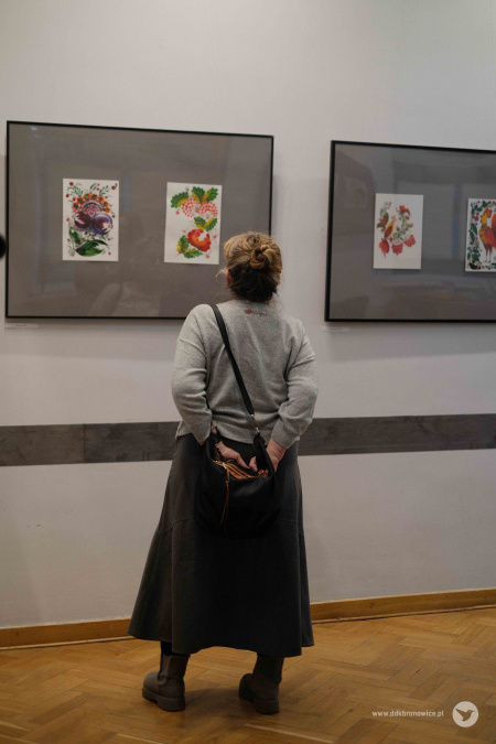 Kobieta w szarym swetrze i w szarej spódnicy ogląda wystawę zawieszonych obrazów. Obrazy wiszą na dwóch szarych tłach.