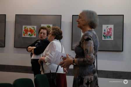 Trzy starsze kobiety oglądają wystawę. Dwie z nich rozmawiają. Jedna z nich ubrana jest w białą bluzkę a druga w czarny sweter. Kobieta na pierwszym planie w szarej bluzce trzyma broszurę wystawową.