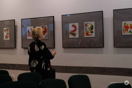 Kobieta w czarno-białej koszuli ogląda wystawę. Obrazy zawieszona są na szarych tłach.