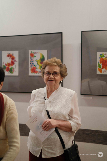 Starsza uczestniczka wystawy. Ubrana jest w białą koszulę. Za nią kolorowe obrazy na białej ścianie.