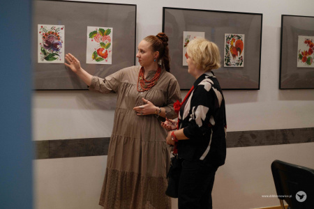 Instruktorka grupy Olena Kovalenko rozmawia z kobietą w czarno-białej koszuli. Instruktorka ubrana jest w bezowa sukienkę i wskazuje na wiszący obraz.