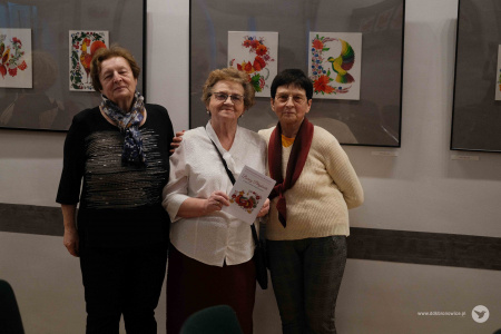 Trzy panie pozują do zdjęcia na tle kolorowych obrazów. Kobieta w środku trzyma w ręce broszurę wystawową.