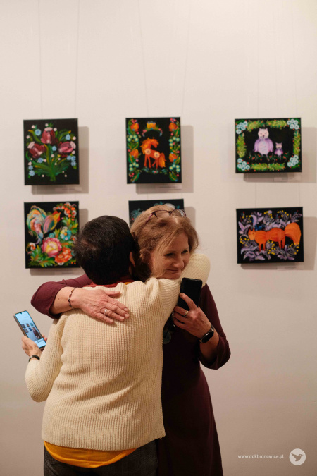 Kobieta w jasnym swetrze ściska Aleksandrę Szulc-Chomę. W tle kolorowe obrazy na białej ścianie.