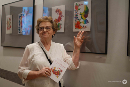 Kobieta w białej koszuli trzyma broszurę wystawową. Wskazuje na swój autorski obraz zawieszony na wystawie. Uśmiecha się.