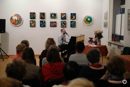 Na pierwszym planie publiczność siedzi na rozstawionych krzesłach. W tle mężczyzna gra na pianinie. Na ścianie zawieszone kolorowe obrazy.