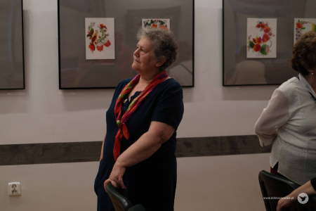 Starsza kobieta w czarnej koszulce i z kolorową chustą na szyi ogląda wystawę. W tle kolorowe obrazy na szarym tle.