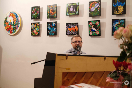 Mężczyzna gra na pinie. Za nim, na białej ścianie, rozwieszone w dwóch rzędach kolorowe obrazy.