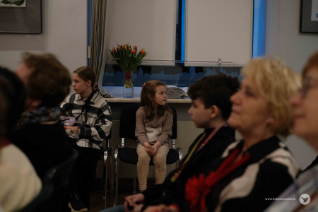 Na pierwszym planie publiczność. W tle, przy ścianie na krzesłach siedzą dwie dziewczynki.