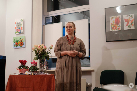 Obok stoliku z kwiatami stoi Olena Kovalenko. Zwrócona jest do widowni. Uśmiecha się.