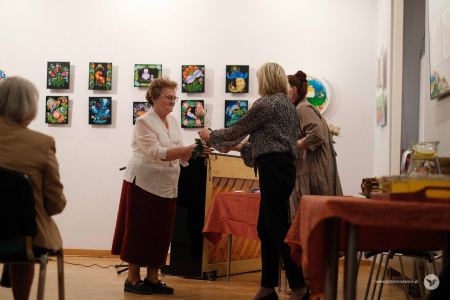 Starsza autorka ubrana w białą koszulę i brązowa spódnicę odbiera kwiaty i dyplom.