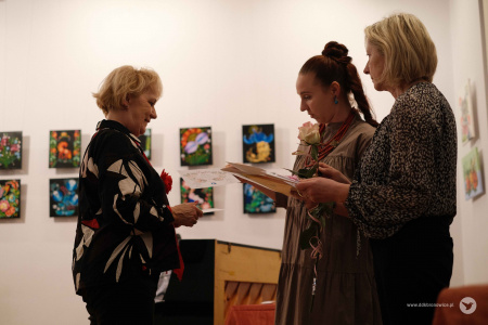 Starsza pani ubrana w biało-czarna koszulę odbiera kwiaty i dyplom z rąk Ewy Rejmak i Oleny Kovalenko.