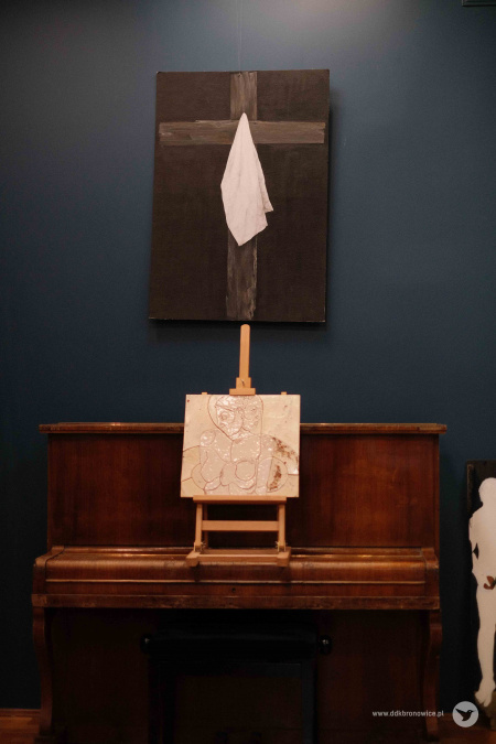 Nad brązowym pianinem wisi obraz. Przedstawia krzyż, na którym jest zawieszona biała chusta. Na pianinie, na sztaludze leży obraz ceramiczny przedstawiający kontury człowieka.